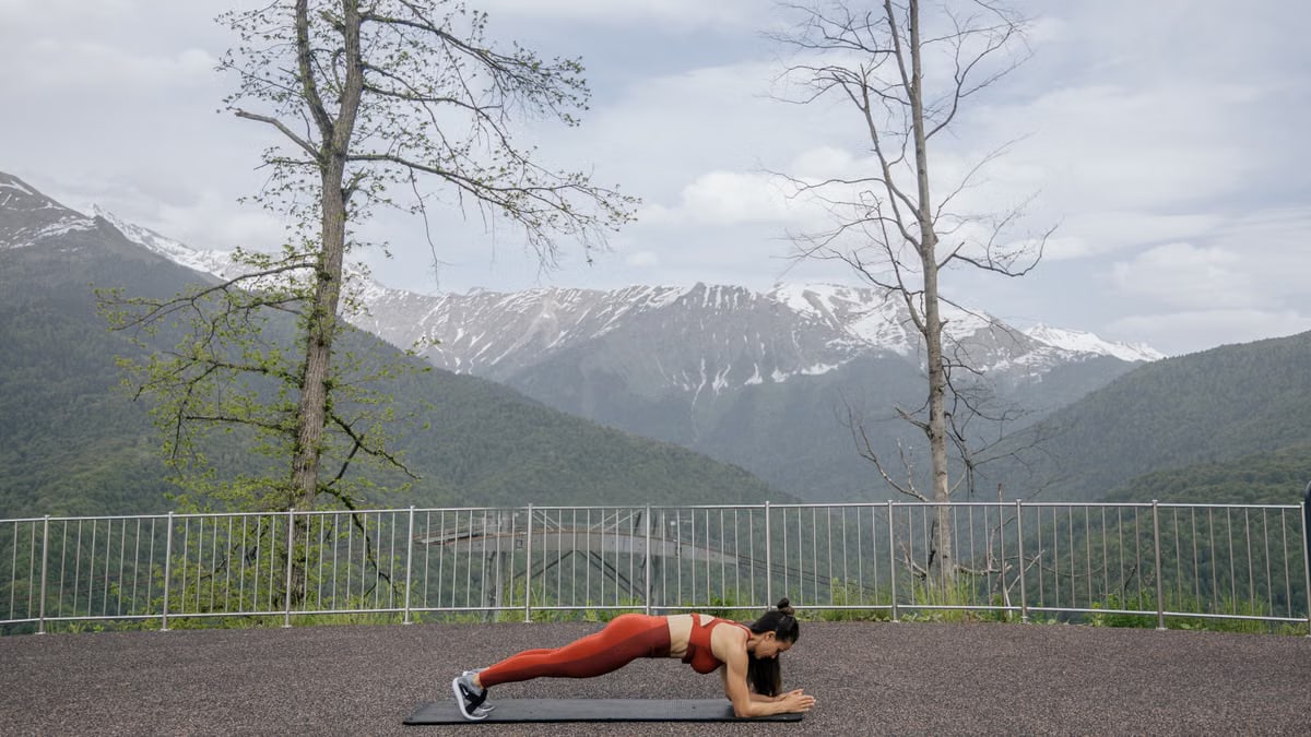 A woman doing a plank exercise on a mat outdoors with a scenic mountain view in the background.