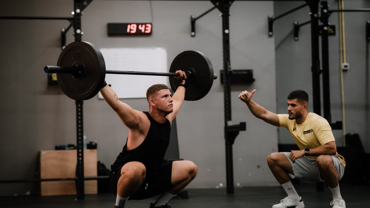 A muscular man performing a weightlifting squat with a coach guiding him indoors.